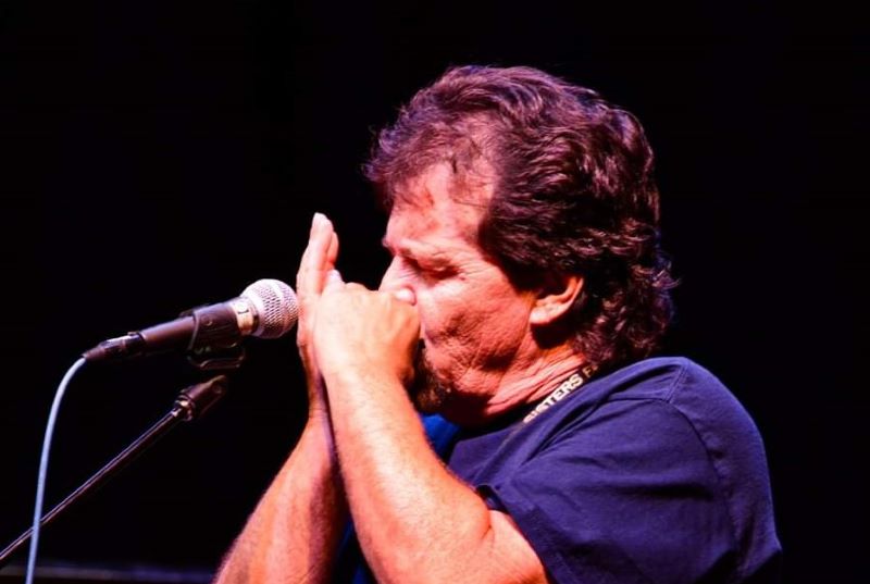 Tim Fast on stage playing harmonica. Wearing Blue Shirt and the venue is dark with lights appearing on Tim. Displays in white text displays in white of Biography of Tim Fast