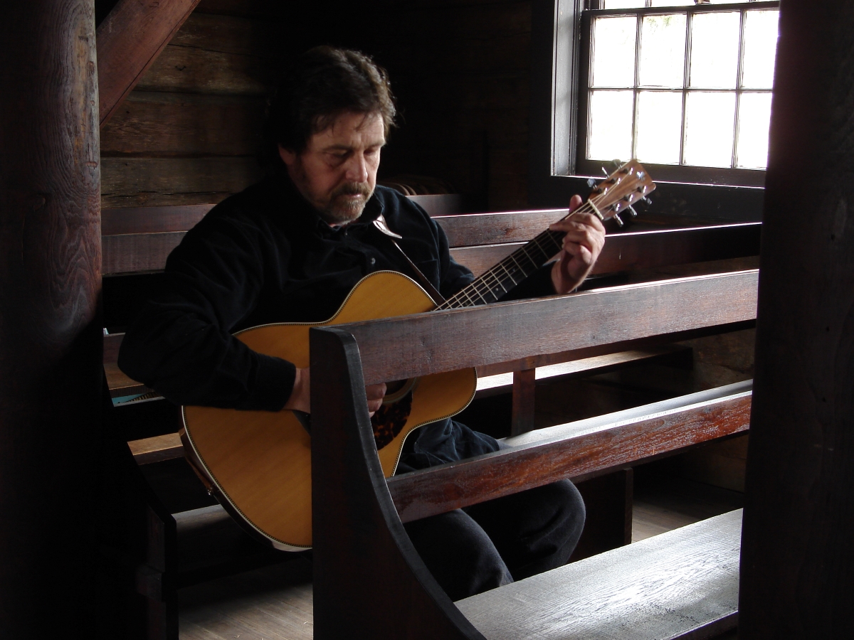 Beautiful historic wood building, Tim Fast sitting and plane acoustic guitar Next to a window with natural light coming in reflecting off of him. Text in white displays. Bookings and General Inquiries